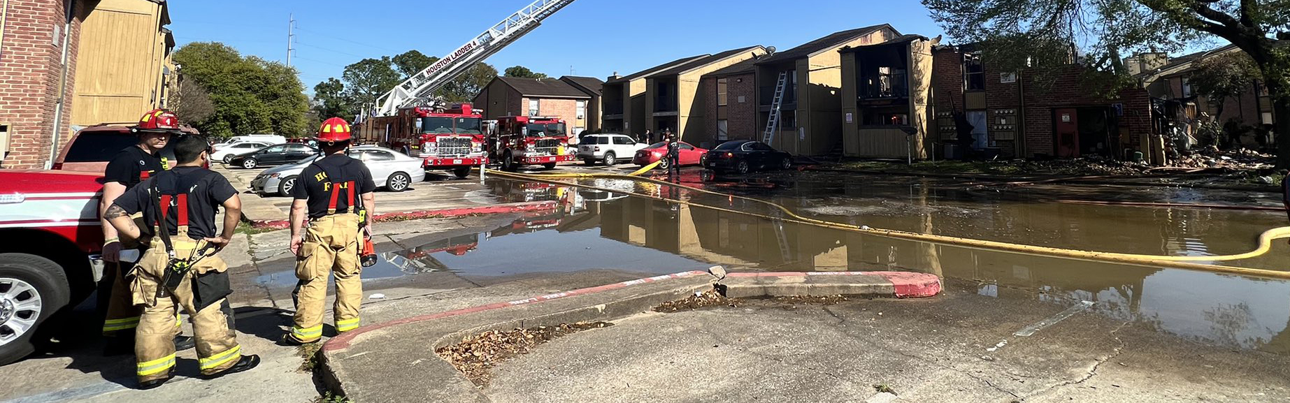 several firefighters stand near an HFD truck looking at an apartment complex with other fire trucks and equipment following a major fire.